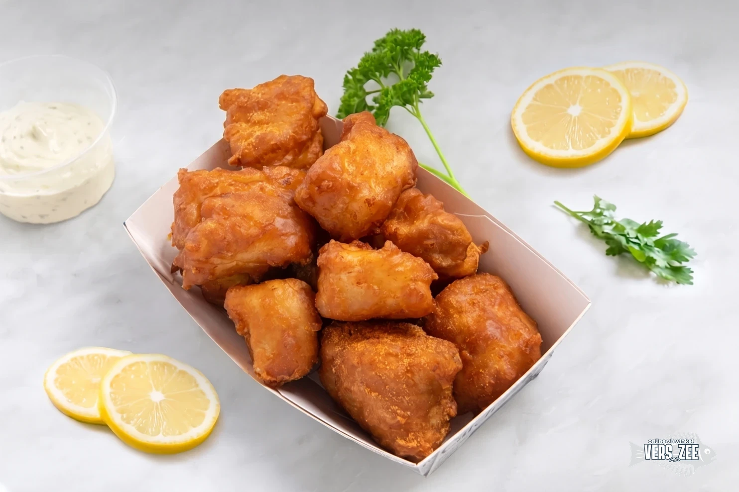 Portie kibbeling (gebakken stukjes vis in beslag) in een bakje, geserveerd met remouladesaus, citroen en peterselie op een lichte marmeren ondergrond.