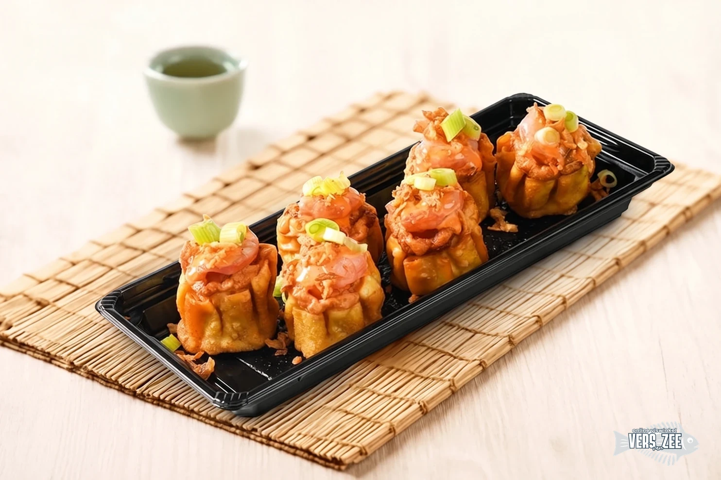 Vijf gefrituurde open dumplings met zalm en garnituur van bosui in zwarte tray, geserveerd op een bamboemat met groene theekop op lichte ondergrond.