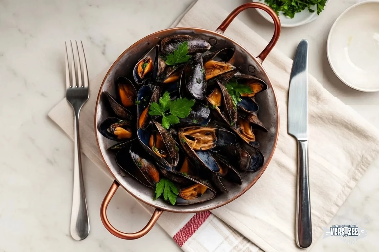 Zwarte mosselen geserveerd in een koperen pan, met verse peterselie, op een wit servet, omgeven door een vork, mes en lege borden op een lichte marmeren ondergrond.